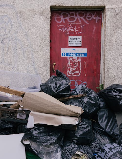 A photograph of a collection of discarded household items placed on a paved driveway outside a residential property. The foreground features a pile of various waste materials, including a large black