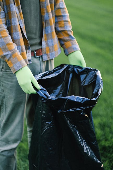 A person wearing a white glove is holding the top of a large white waste bag filled with a mixture of garden debris, including small green leaves, twigs, and dried foliage. A small shovel with a weath