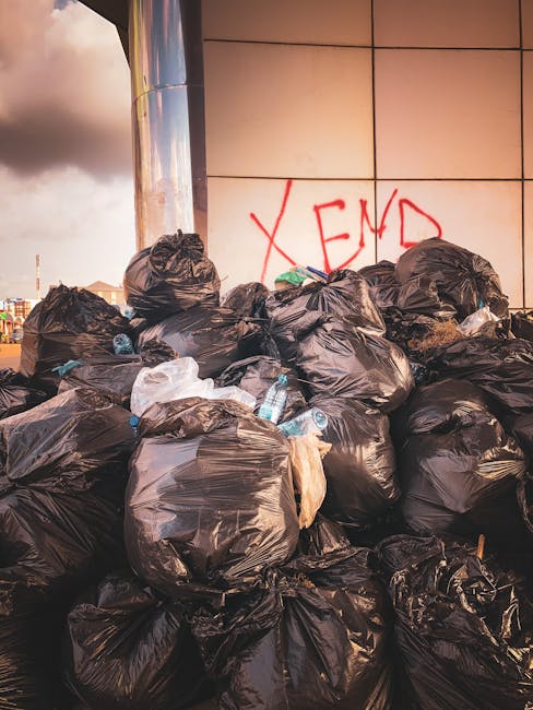 A large pile of black plastic rubbish bags is stacked against a tiled wall, with some of the bags appearing to be filled with general waste or household refuse. Several clear plastic bottles, partiall